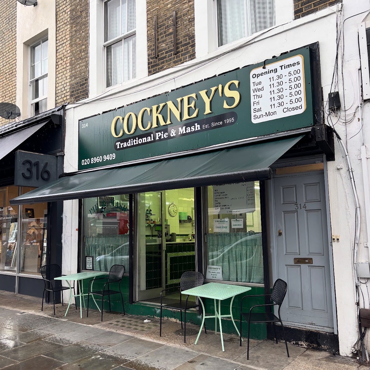 The shopfront at 314 Portobello Road: a dark green fascia board with COCKNEY'S in gold shadowed capitals, 'Traditional Pie & Mash' and 'Est. Since 1995' beneath it, a green awning, green gingham half-curtains in the window, and mint-green metal tables on the wet pavement outside.