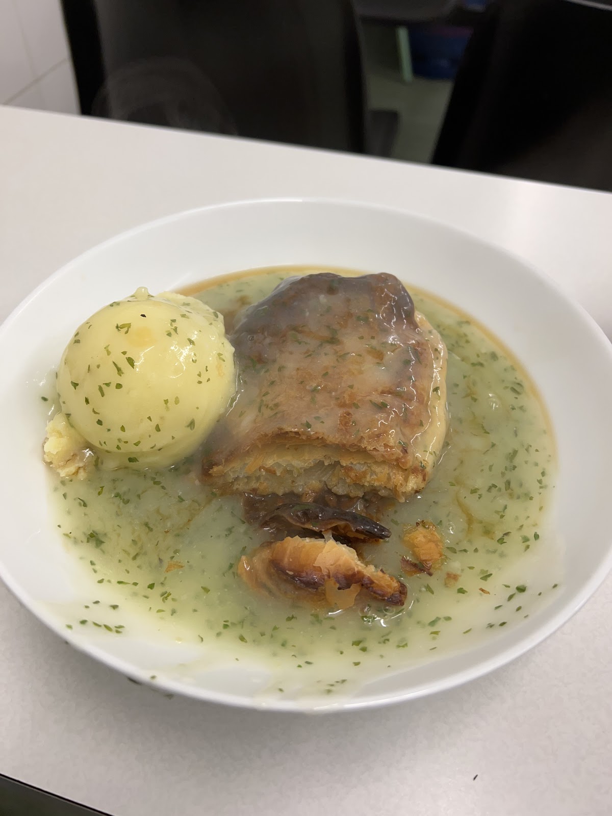 A pie broken open beside a round scoop of mashed potato in a white bowl of parsley liquor, on a white counter.