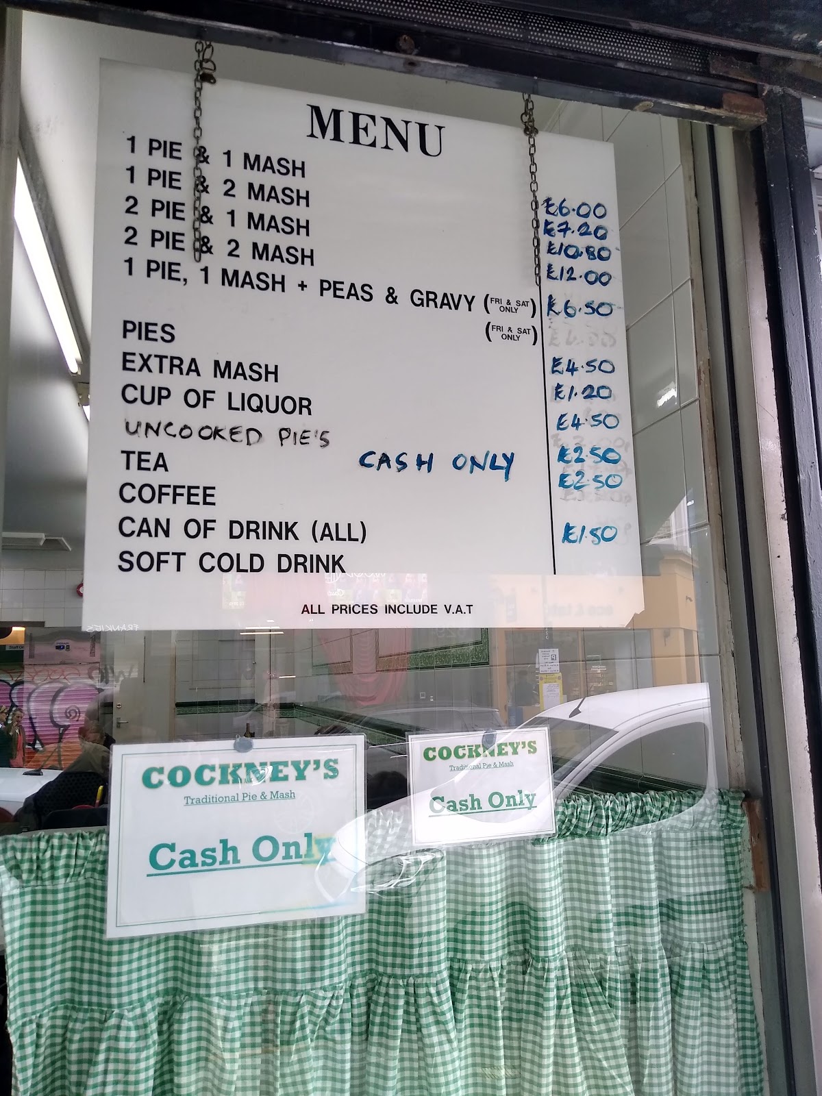 The price board hanging in the shop window: a white sign headed MENU listing pie and mash combinations with handwritten blue prices, the words CASH ONLY written across it, and below it two printed COCKNEY'S Traditional Pie & Mash 'Cash Only' notices above green gingham half-curtains.