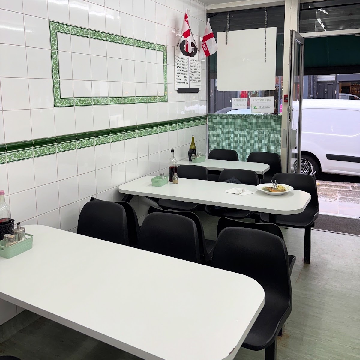 Inside the shop: white tiled walls with a green patterned border, white formica tables with black chairs, vinegar and chilli vinegar bottles in a mint-green caddy, and green gingham curtains at the window.