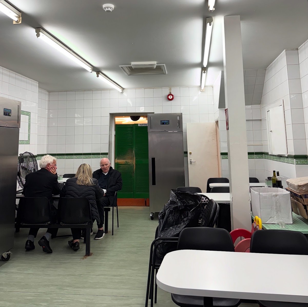 The back dining room: three customers at a table under strip lighting, white tiling with a green border, a green louvred Staff Only door and stainless steel fridges.